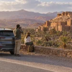 Voiture de location sur une route asphaltée près d’une kasbah de Ouarzazate, avec des montagnes arides au loin.