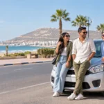 Trois jeunes amis souriants près d’une voiture compacte sur le front de mer d’Agadir